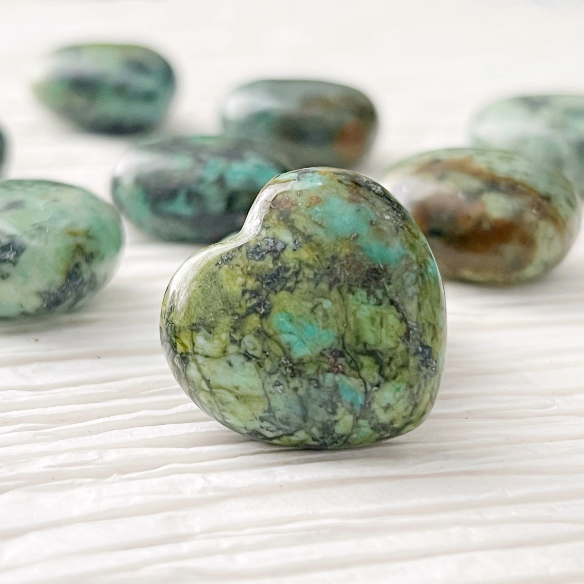 1" Mini African Turquoise Hearts are displayed, with some resting on a white, round, translucent surface and others on a gold dish. Green foliage is partially visible in the background. The heart-shaped gemstones feature a mix of green and darker shades, creating a natural pattern.