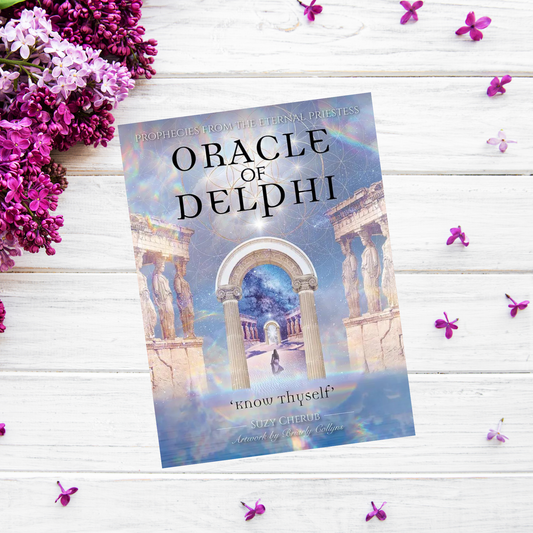 A book titled "Oracle Of Delphi: Prophecies from the Eternal Priestess" is placed on a white wooden surface surrounded by scattered small purple flowers and a bunch of purple flowers on the left. The book's cover, reminiscent of an oracle deck, features ancient Greek ruins under a mystical sky.