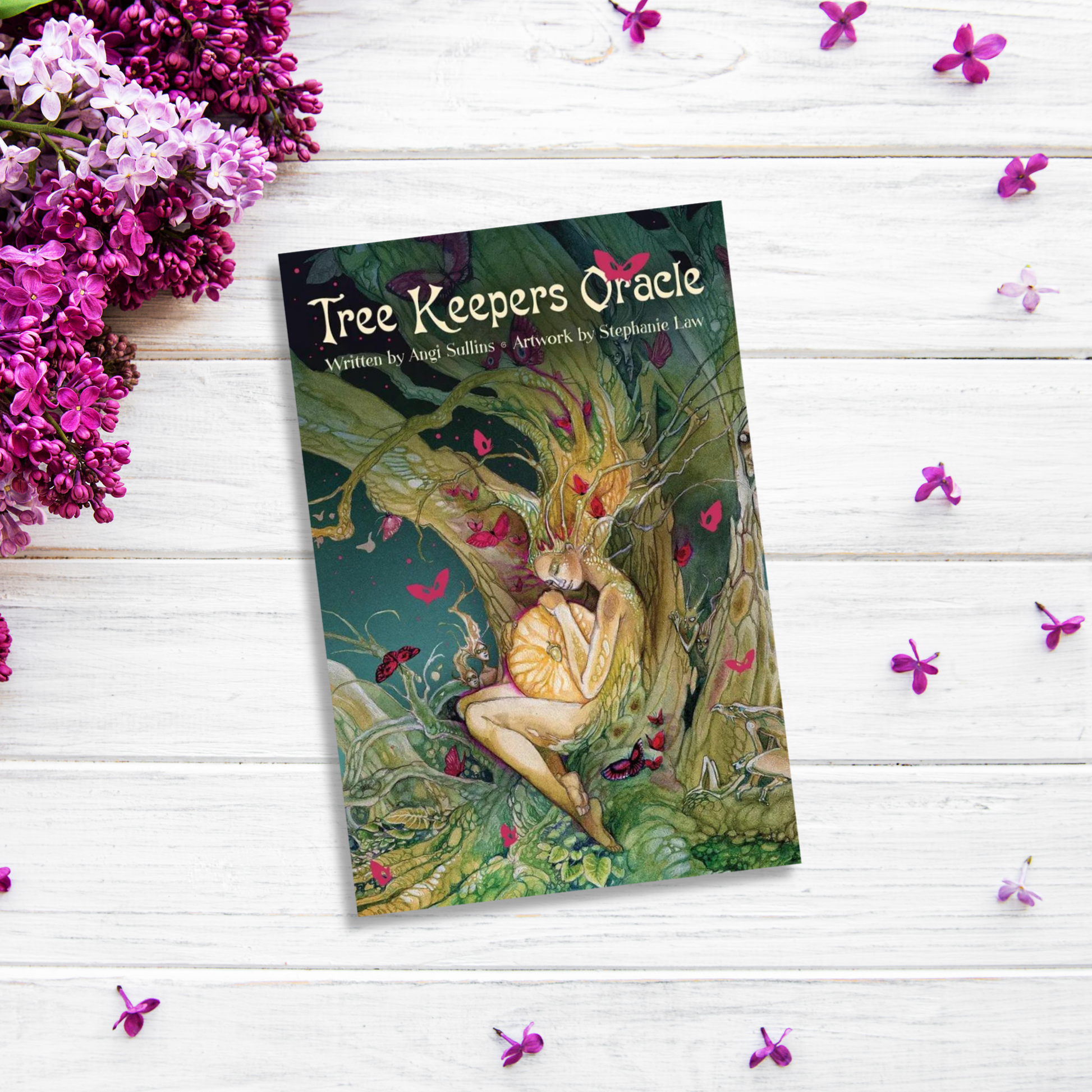 A book titled "Tree Keepers Oracle," with illustrations by Stephanie Law, lies on a white wooden surface, surrounded by lilac flowers. The cover art depicts a person cradling a glowing orb while sitting against an intricate, ancient tree, inviting readers into magical worlds.