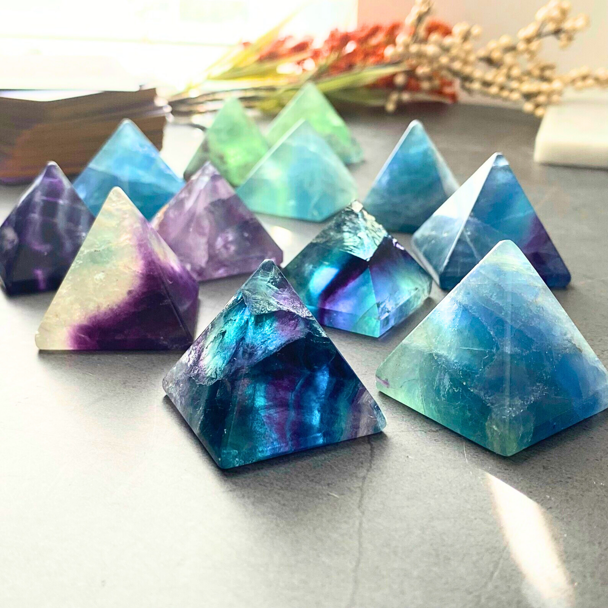 A hand with sparkly pink nail polish holds a transparent 1" Mini Rainbow Fluorite Pyramid with shades of blue, purple, and green. The background is an outdoor setting with soft-focus greenery and a light-colored building, perfect for mental and spiritual growth or to boost creativity.