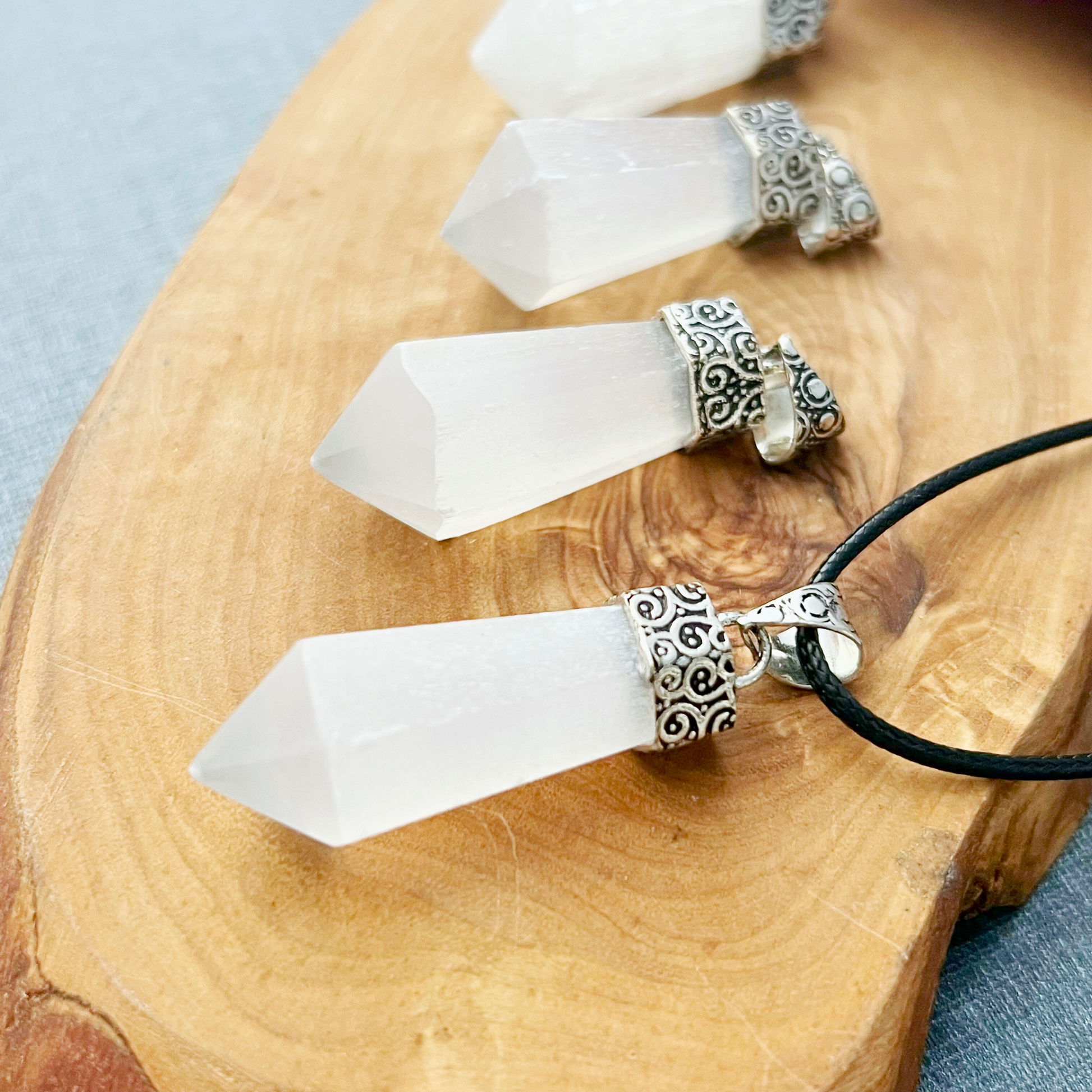 A close-up of three white 1.5" Selenite Point Pendants with ornate antique silver caps, arranged on a wooden surface. One pendant, known for its metaphysical properties, is attached to a black cord and ready to wear as a necklace. The crystals are cut into pointed, hexagonal shapes.