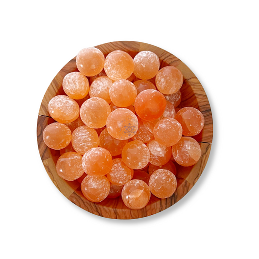 A top view of a wooden bowl filled with numerous round, orange candies resembling Orange Selenite Tumbled Sphere Stones. The candies have a slightly translucent appearance, with a sugary coating visible on their surfaces, invoking thoughts of emotional well-being. The bowl is set against a white background.