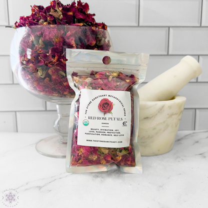 A sealed packet labeled "Red Rose Petals" stands on a marble surface, next to a clear glass bowl filled with loose dried rose petals perfect for ritual baths. A white marble mortar and pestle are on the right, set against a white tile background.