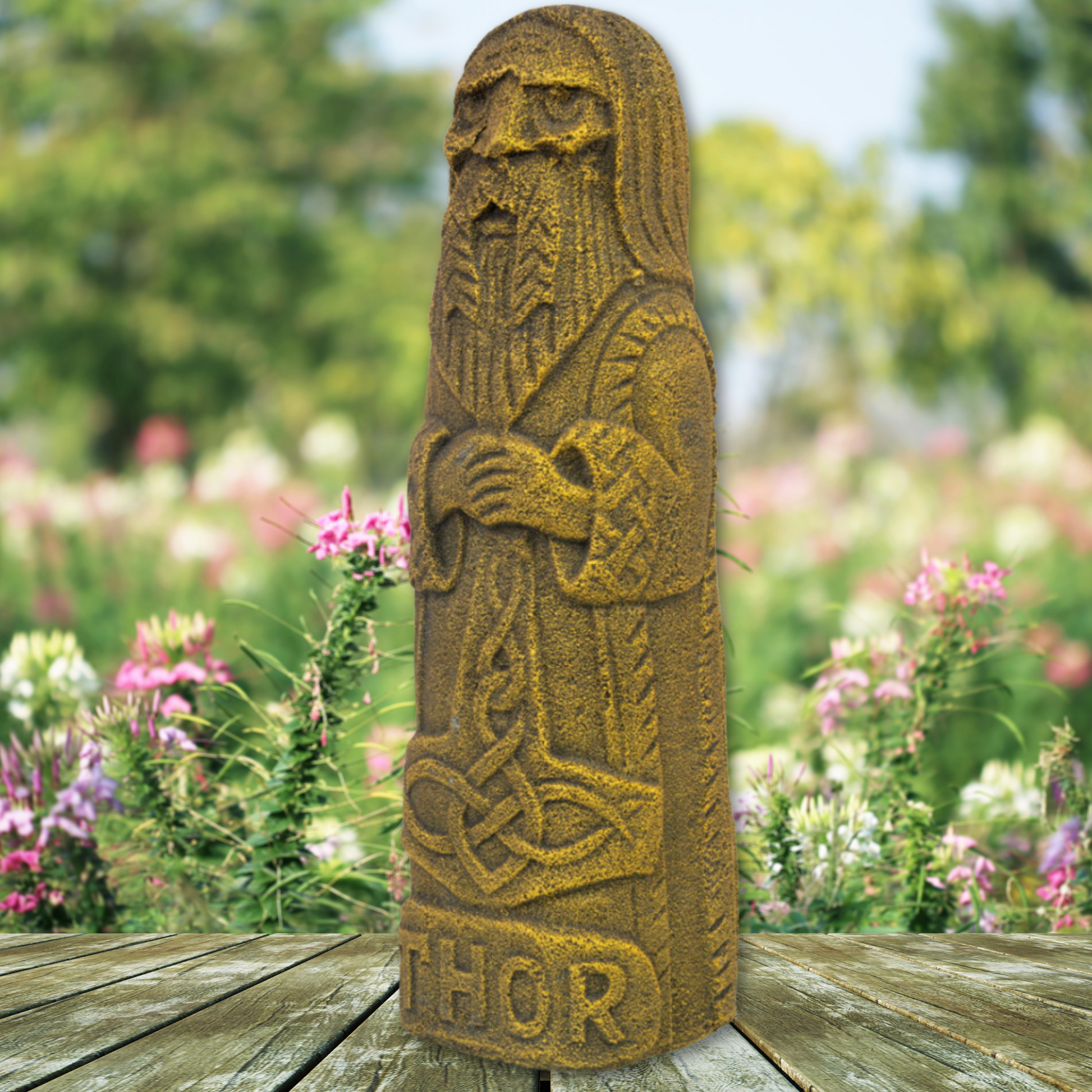 A weathered statue of a bearded figure wearing a robe, hands clasped at the chest, stands on a wooden platform. This Thor Volcanic Statue has "THOR" engraved at the base and is set against a blurred background of blooming wildflowers and greenery.