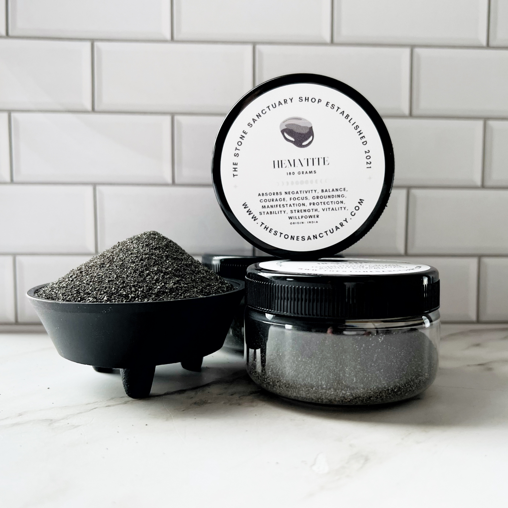 A close-up image of three jars labeled "Hematite Gemstone Sand" against a white tile background. One jar is open, displaying a small pile of black, fine-grained natural Hematite Gemstone Sand in a round container. The jars are from "The Stone Sanctuary Shop.