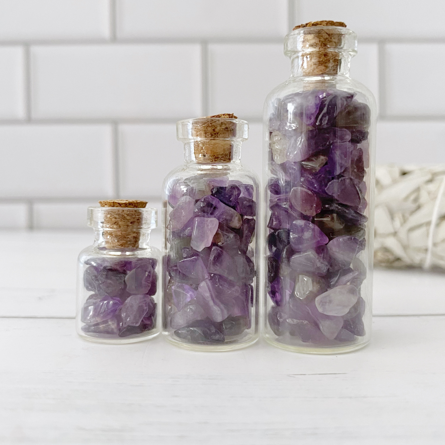 A wooden bowl filled with small polished Amethyst Crystal Chips is placed on a white surface. Surrounding the bowl are scattered amethyst stones, small bottles containing crystals, and a bundle of white sage to enhance psychic abilities. A plastic bag of stones is also visible.