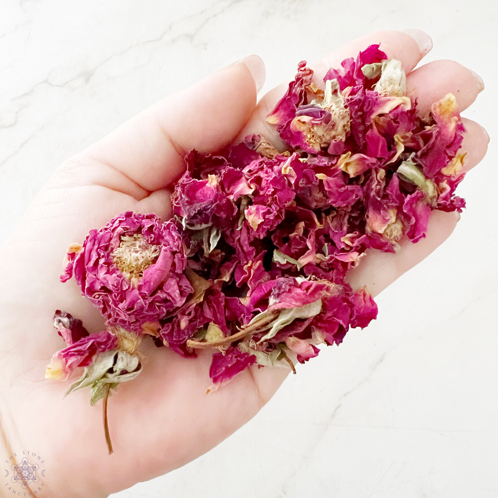 A sealed packet labeled "Red Rose Petals" stands on a marble surface, next to a clear glass bowl filled with loose dried rose petals perfect for ritual baths. A white marble mortar and pestle are on the right, set against a white tile background.