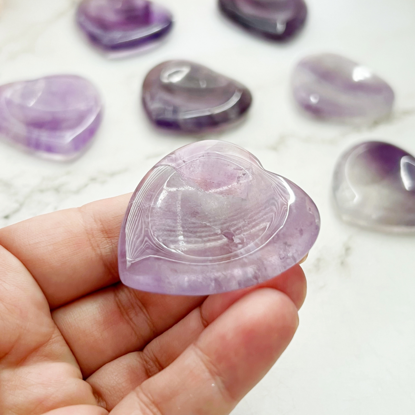 A wooden tray topped with a pink flower, green leaf, and five polished Amethyst Heart Shaped Worry Stones in varying shades of purple rests on a white marble surface, promoting stress relief and emotional balance.