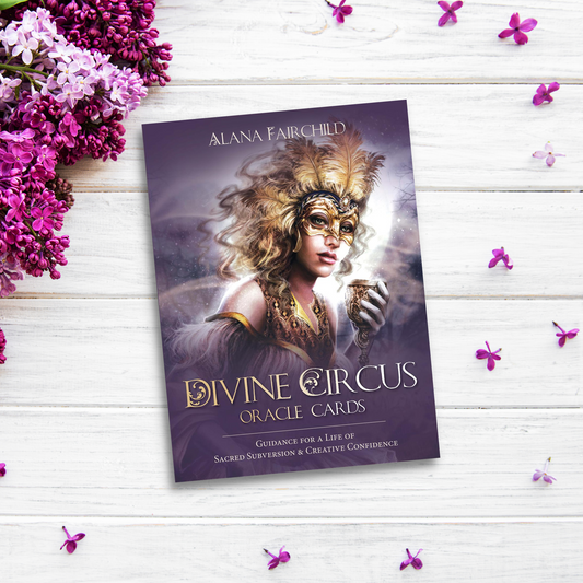 A "Divine Circus Oracle Cards" deck by Alana Fairchild rests on a white wooden surface surrounded by purple flowers. The cover depicts a mystical character in a mask holding a cup, set against a cosmic background. Text on the cover reads: "Guidance for Bold-Spirited Individuals & Creative Confidence.