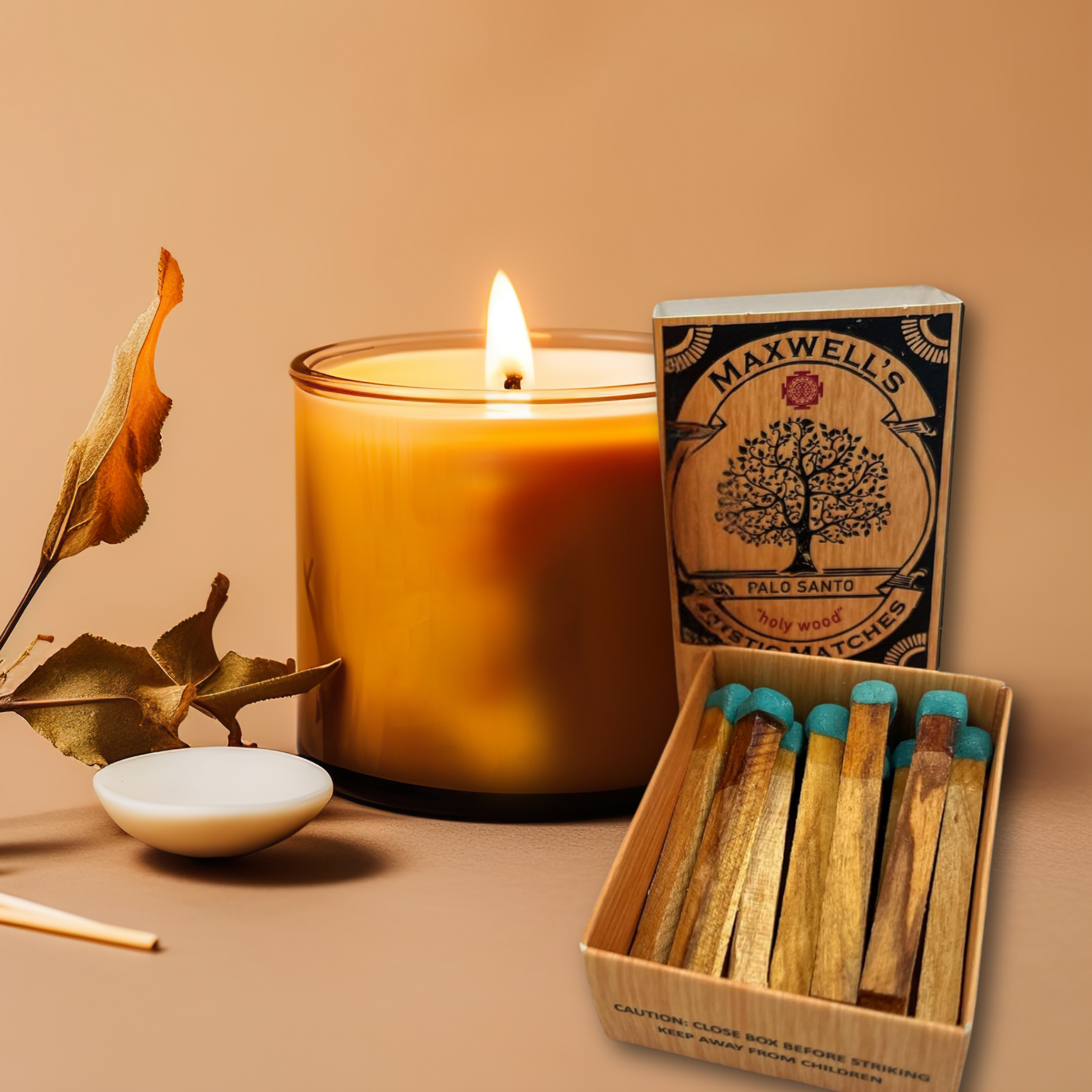 A lit amber candle sits on a beige surface. Next to it lies a wooden matchbox labeled "Maxwell's Mystic Palo Santo Matches," ideal for cleansing rituals and spiritual practice, with several matches inside. A small white dish holds a single match, while a dried leaf rests to the left. The background is beige.