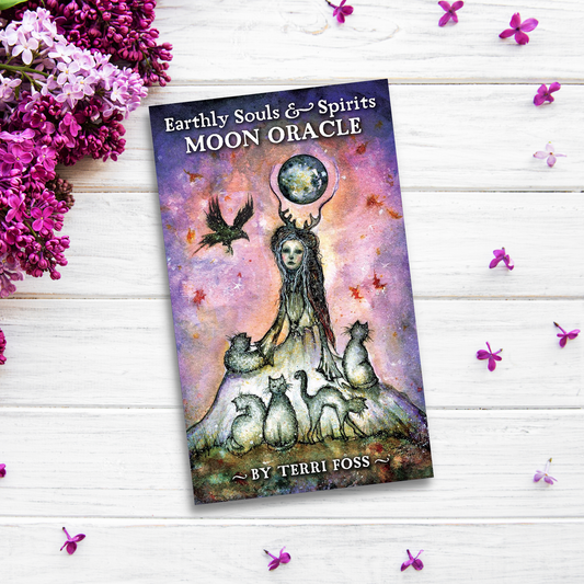 The image shows the Earthly Souls & Spirits Moon Oracle deck by Terri Foss placed on a white wooden surface, surrounded by pink and purple flowers. The card's artwork features a mystical figure holding a glowing orb with animals and a bird around them, emanating intense spiritual energy.