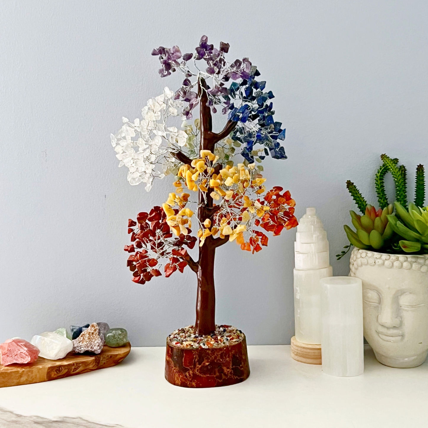 A 15" Seven Chakra Gemstone Tree with branches adorned with multicolored crystal leaves stands on a table. The tree features white, purple, blue, yellow, and orange leaves representing the seven chakras. Nearby are various crystals and succulent plants, including a Buddha head planter and a white crystal tower.