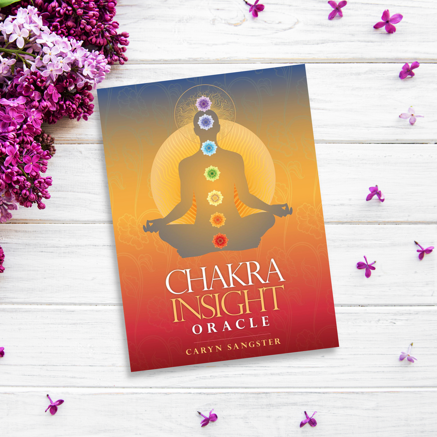 A book titled "Chakra Insight Oracle" by Caryn Sangster lies on a white wooden surface surrounded by purple flowers. This Chakra Insight Oracle's cover features a silhouette of a meditating figure with chakras aligned against a gradient background from blue at the top to orange at the bottom.