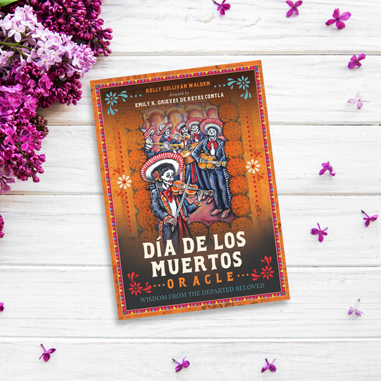 A book titled "Día De Los Muertos Oracle" lies on a white wooden surface decorated with purple flowers. The cover features colorful illustrations of skeletons in traditional Mexican attire, playing musical instruments and celebrating the Day of the Dead, offering deep ancestral wisdom.