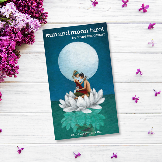 A tarot deck box titled "Sun and Moon Tarot" is displayed. The illustration shows a couple sitting closely together on a large lotus flower under a full moon against a blue background. The box rests on a white wooden surface adorned with scattered purple and white flowers, blending elegance with mysticism.