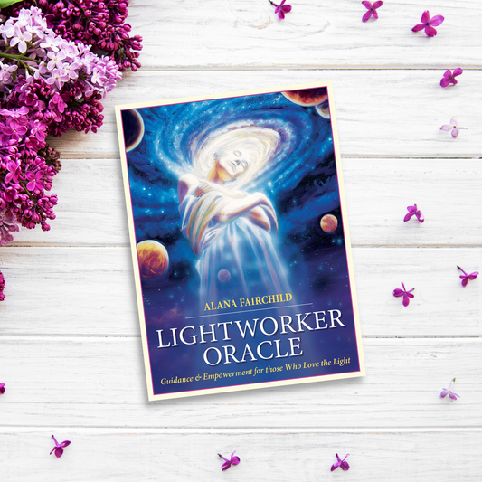 A book titled "Lightworker Oracle" by Alana Fairchild rests on a white wooden surface surrounded by purple flowers. The cover features an ethereal figure wrapped in divine light with celestial elements like planets in the background, inviting readers on a profound spiritual journey.