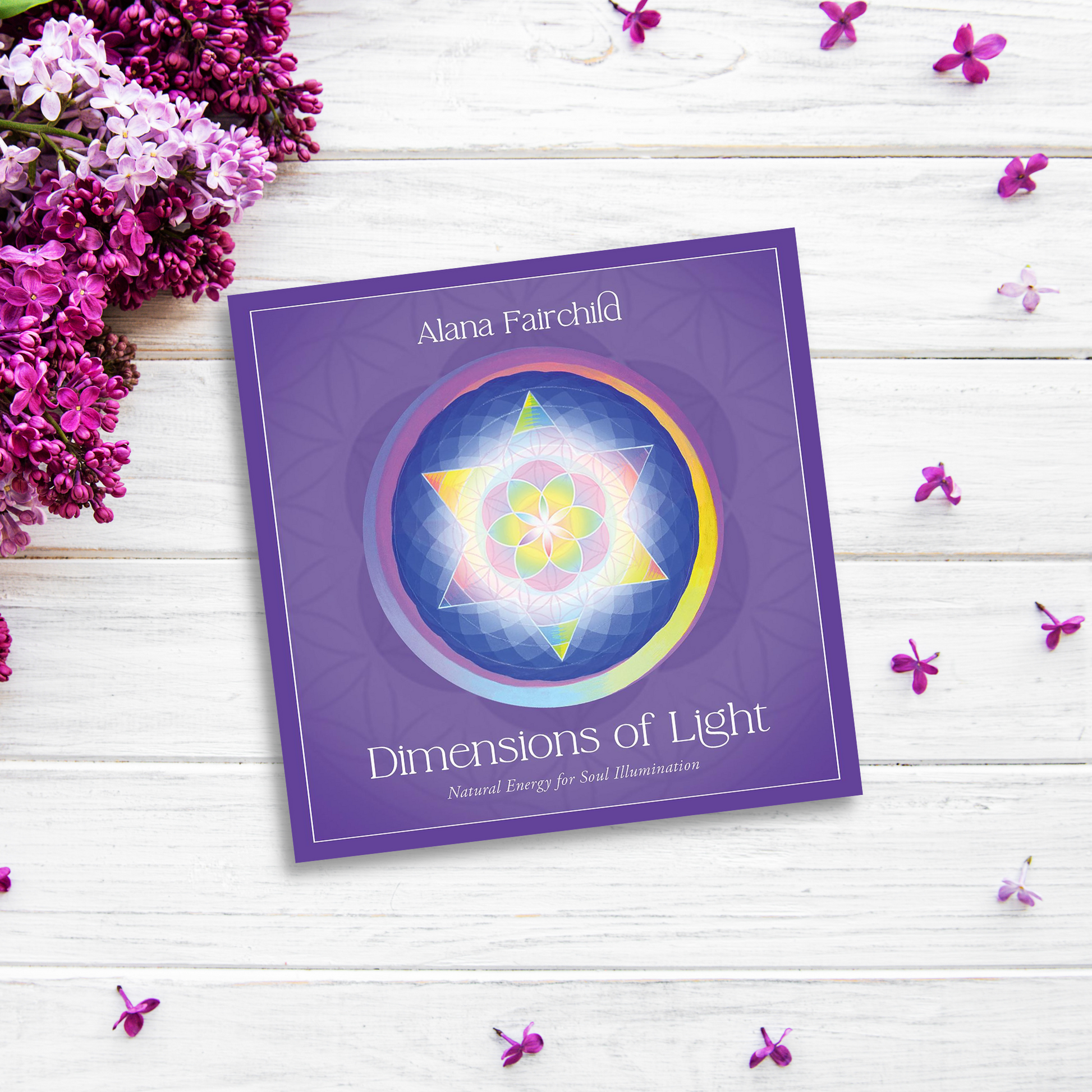 A product titled "Dimensions of Light" by Alana Fairchild lies on a white wooden surface, surrounded by purple flowers and petals. The cover, showcasing vibrant, hand-painted mandalas on a purple background, beautifully complements its theme.