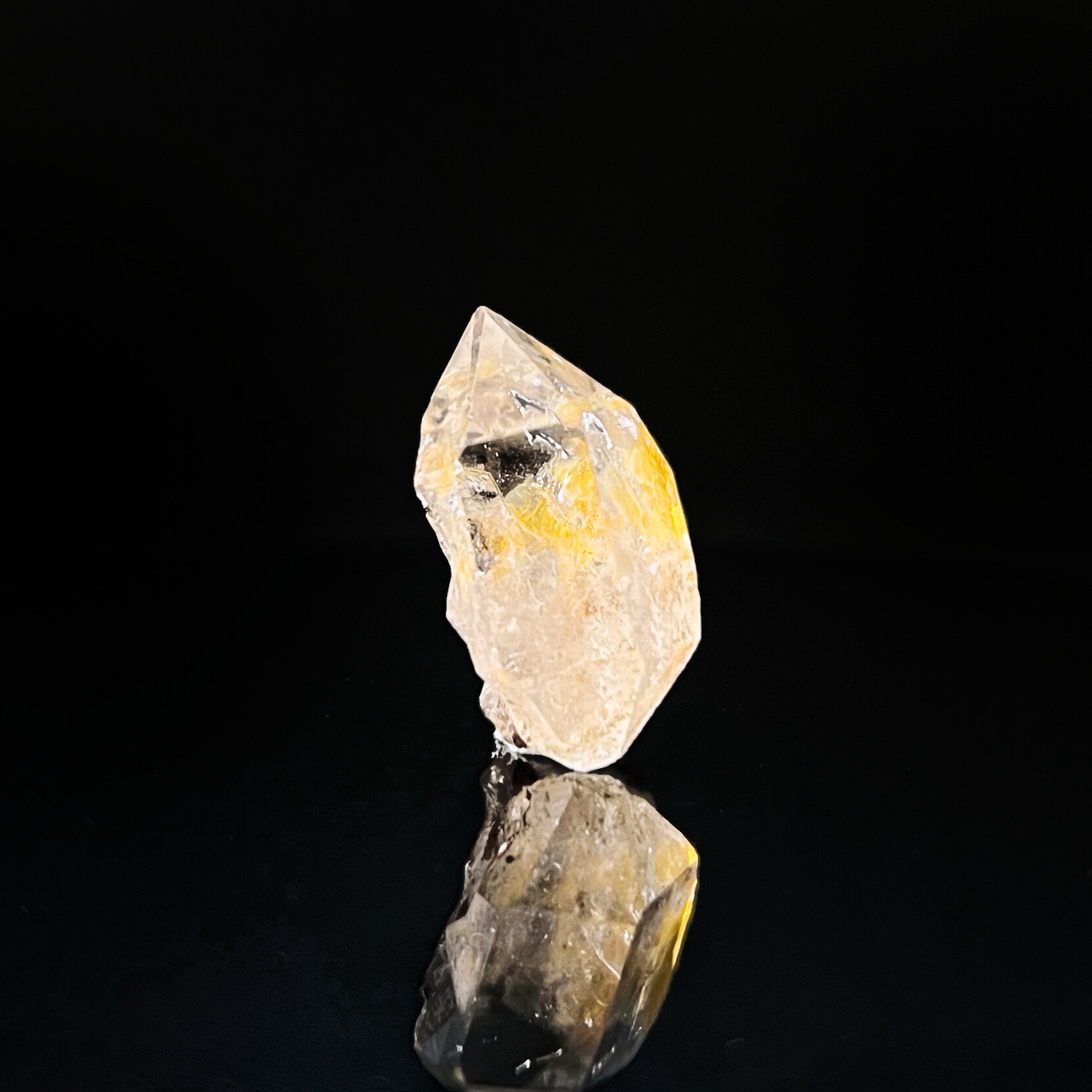 A 1" Raw Herkimer Diamond stands against a black background. The Grade A+ crystal has a slightly irregular shape and features internal fractures and inclusions, with a subtle hint of yellow hues reflecting off its surfaces. Its reflection is visible on the glossy surface below.