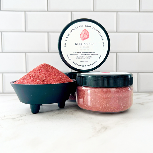 A small black bowl filled with Red Jasper Gemstone Sand, accompanied by two jars of Red Jasper Gemstone Sand on a white tiled surface. The open jar displays a lid labeled "Red Jasper" and a description of its properties, radiating peaceful energy.