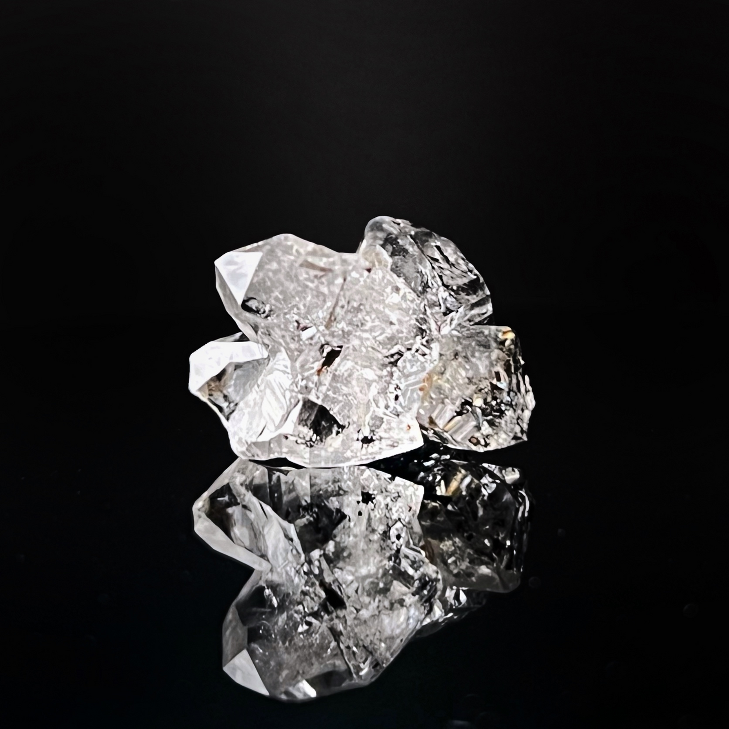 A close-up image of three clear, reflective, irregularly-shaped crystals placed on a glossy black surface, creating a striking reflection below them. The dark background enhances the brilliance and clarity of this .72" Raw Herkimer Diamond Cluster with Hydrocarbon Anthraxolite, revealing its metaphysical properties.