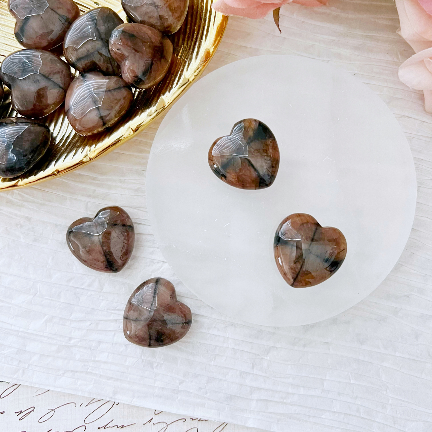 A collection of heart-shaped stones featuring brown and black patterns are neatly arranged on a golden plate and a white circular stone slab. Among them, the 1" Mini Chiastolite Heart stands out, celebrated for its grounding energies. Additional stones and some pink flowers are positioned nearby on a white textured surface.