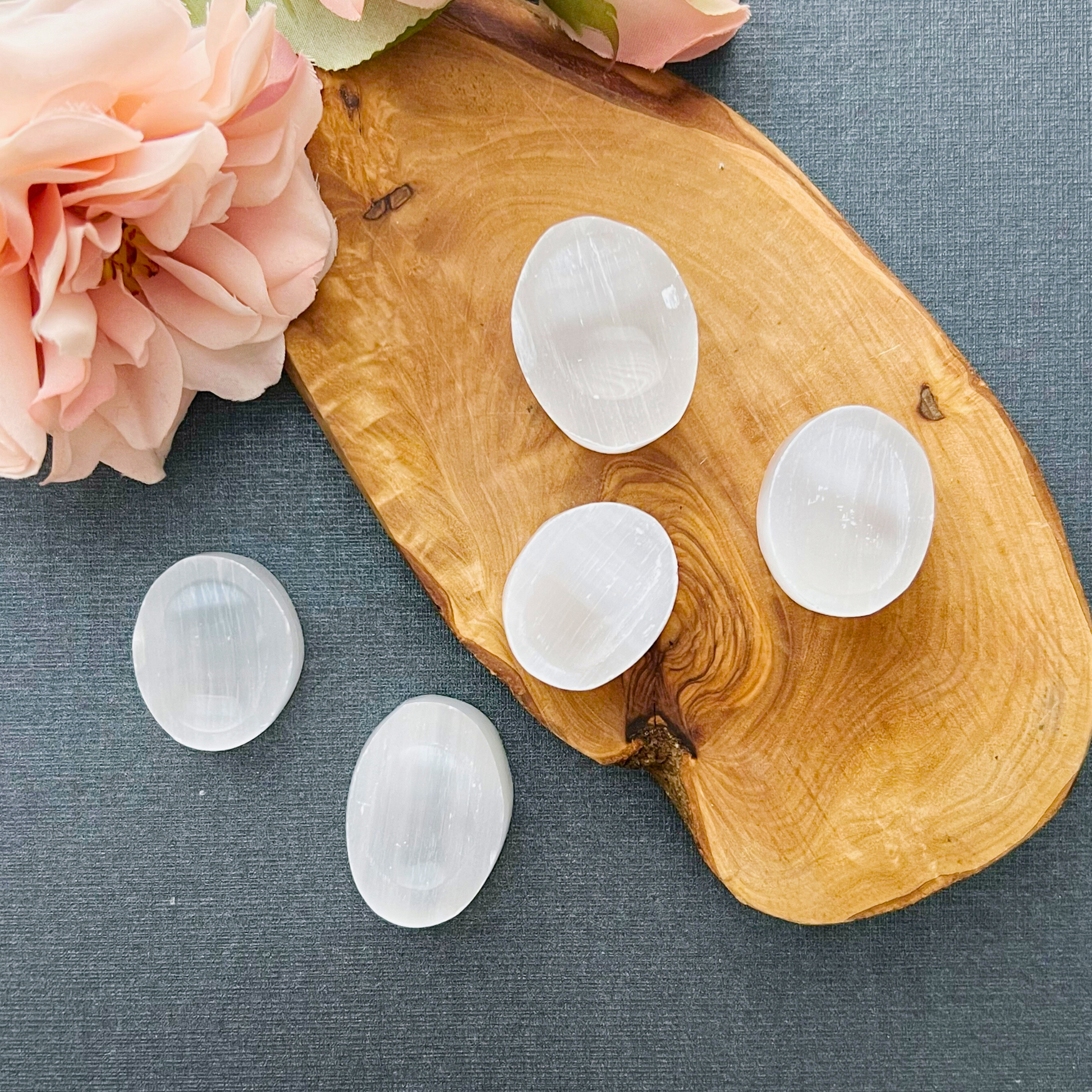 Five 1" Selenite Worry Stones, polished and translucent white, are elegantly arranged on a light wooden slab. The slab rests on a dark, textured surface. A pink flower in the top left corner adds a soft touch to this calming and healing scene.