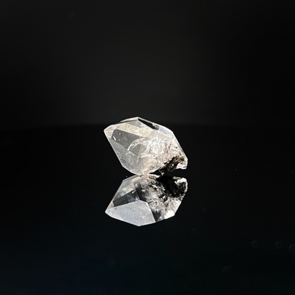 A .63" Raw Herkimer Diamond placed on a reflective black surface creates a mirrored image. The natural quartz crystal catches light, showcasing its translucence and intricate cuts. The background is entirely black, highlighting the brilliance and clarity of these unique formations.