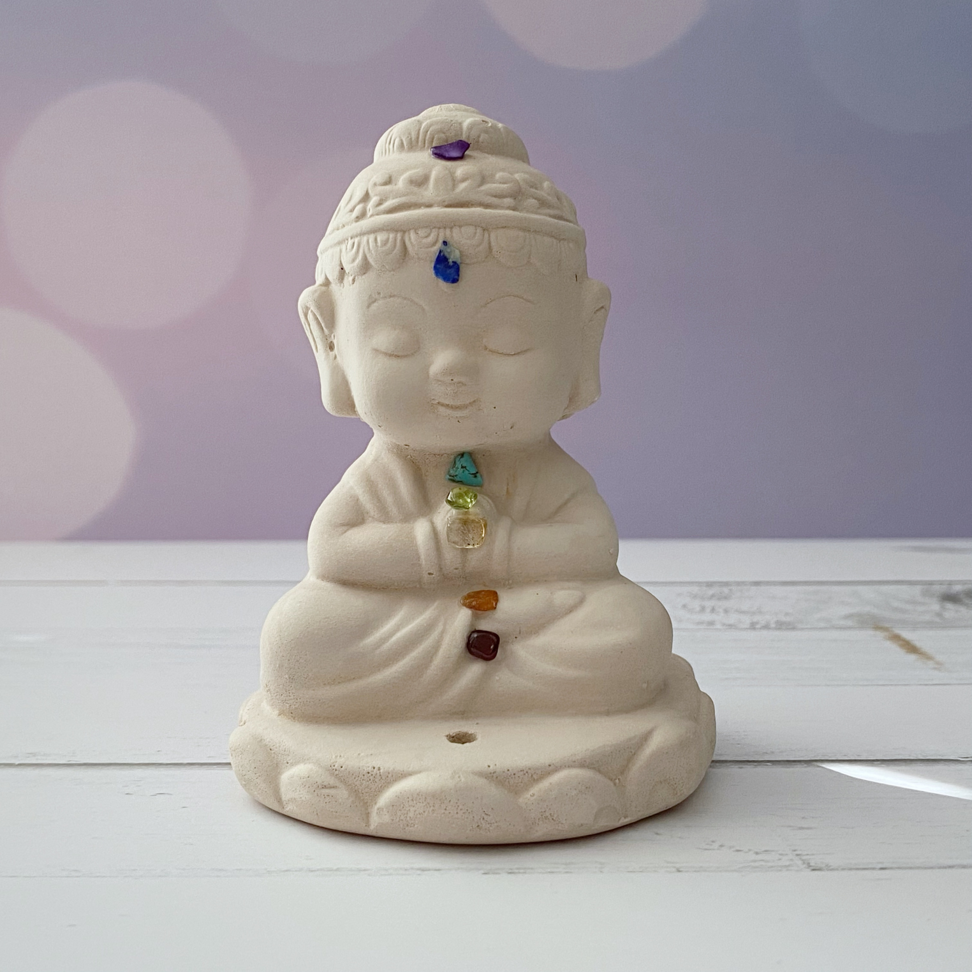 A small ceramic Buddha statue in a seated meditation pose doubles as a Chakra Buddha Incense Holder. The Chakra Buddha Incense Holder features colorful gemstone stone chips on its chakras, representing various energy points. The background is softly blurred with pastel colors and bokeh light effects, set on a white wooden surface.