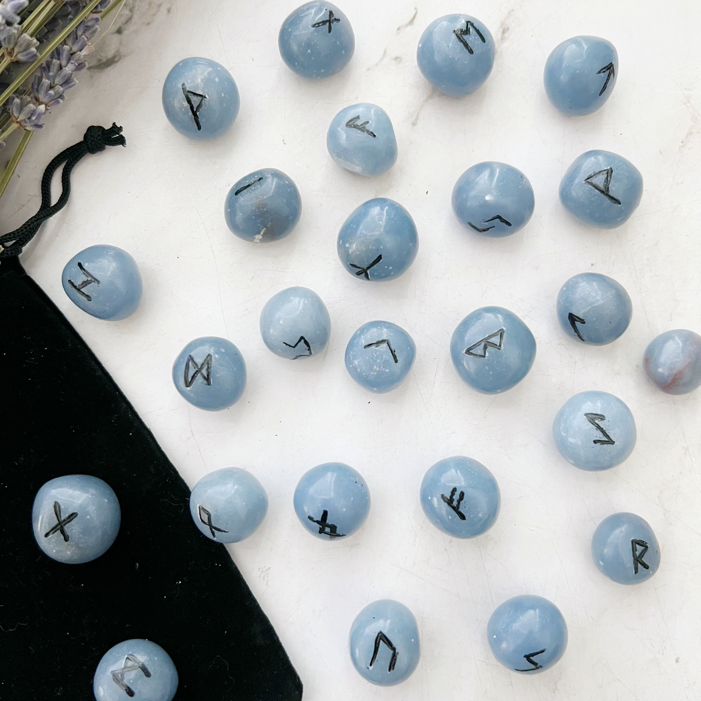 Smooth, blue Angelite Elder Futhark Runes with black Elder Futhark script symbols etched on them are spread out on a white surface. A black drawstring pouch is visible in the bottom left corner. Lavender flowers are partially visible in the top left corner, hinting at their metaphysical properties.