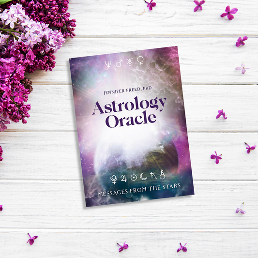 A book titled "Astrology Oracle" by Jennifer Freed, PhD, lies on a white wooden surface surrounded by scattered purple flowers and greenery. This guidebook features a cosmic background with planets, zodiac signs, and imparts astrological wisdom through 64 cards.