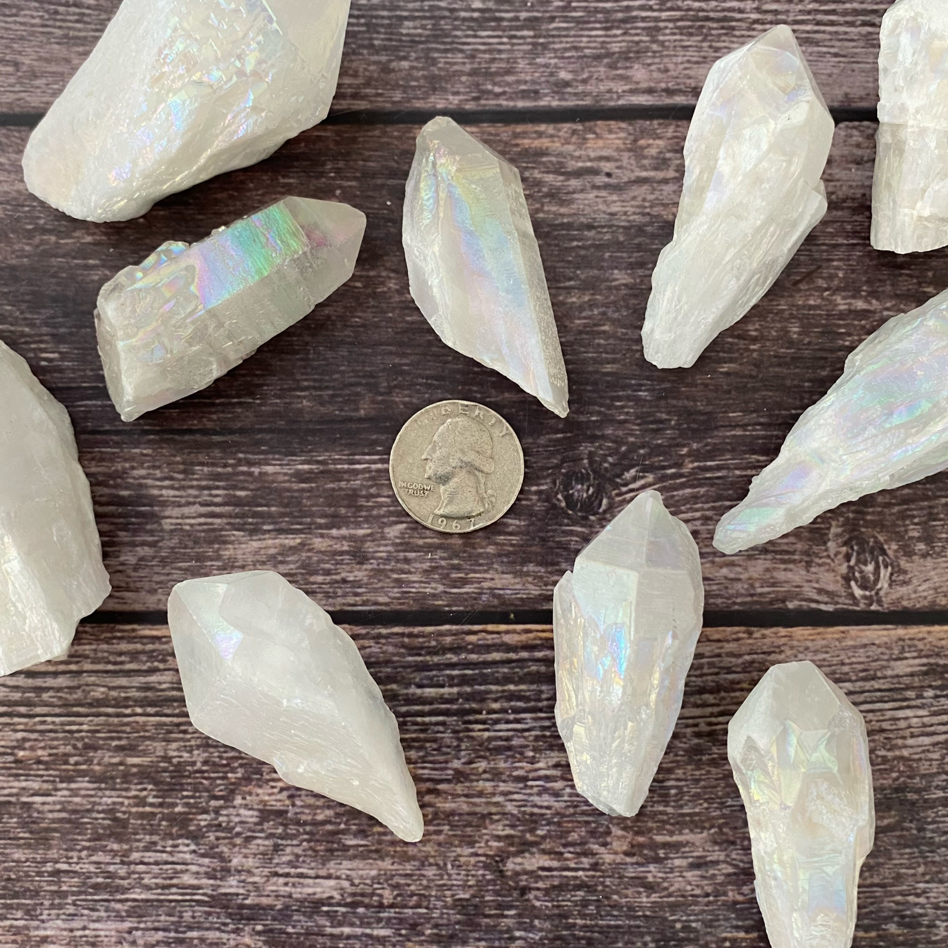 A hand with glittery nail polish holds a large, iridescent 1-4" Raw Angel Aura Quartz Point with a translucent appearance, reflecting rainbow hues. In the background, more similar crystals are displayed in a wooden bowl on a wooden surface, with green foliage partially visible.