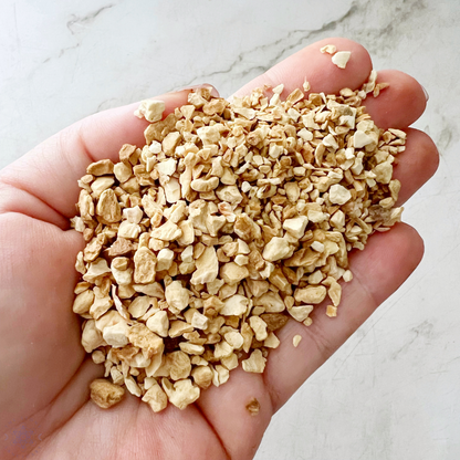 A small, clear resealable bag labeled "Orris Root" is filled with small dried granules. Behind it, a large circular container holds more Orris Root, while next to it rests a white marble mortar and pestle—essential tools for those incorporating Orris Root into their spiritual rituals. The background features a white tiled wall.