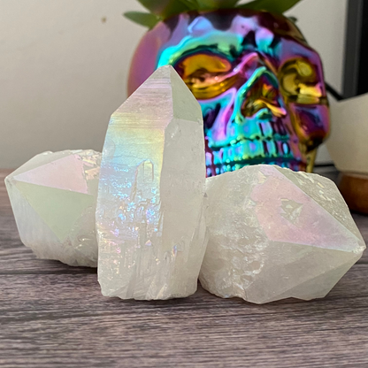 A hand with glittery nail polish holds a large, iridescent 1-4" Raw Angel Aura Quartz Point with a translucent appearance, reflecting rainbow hues. In the background, more similar crystals are displayed in a wooden bowl on a wooden surface, with green foliage partially visible.