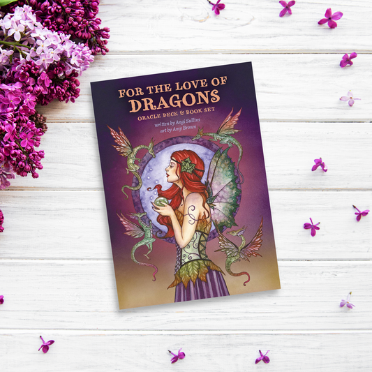 A collector's book titled "For the Love of Dragons: Oracle Deck & Book Set" lies on a white wooden surface. The cover art, possibly illustrated by Amy Brown, depicts a woman in a red headpiece holding a small dragon, surrounded by larger dragons. Purple flowers and petals are scattered around the book.