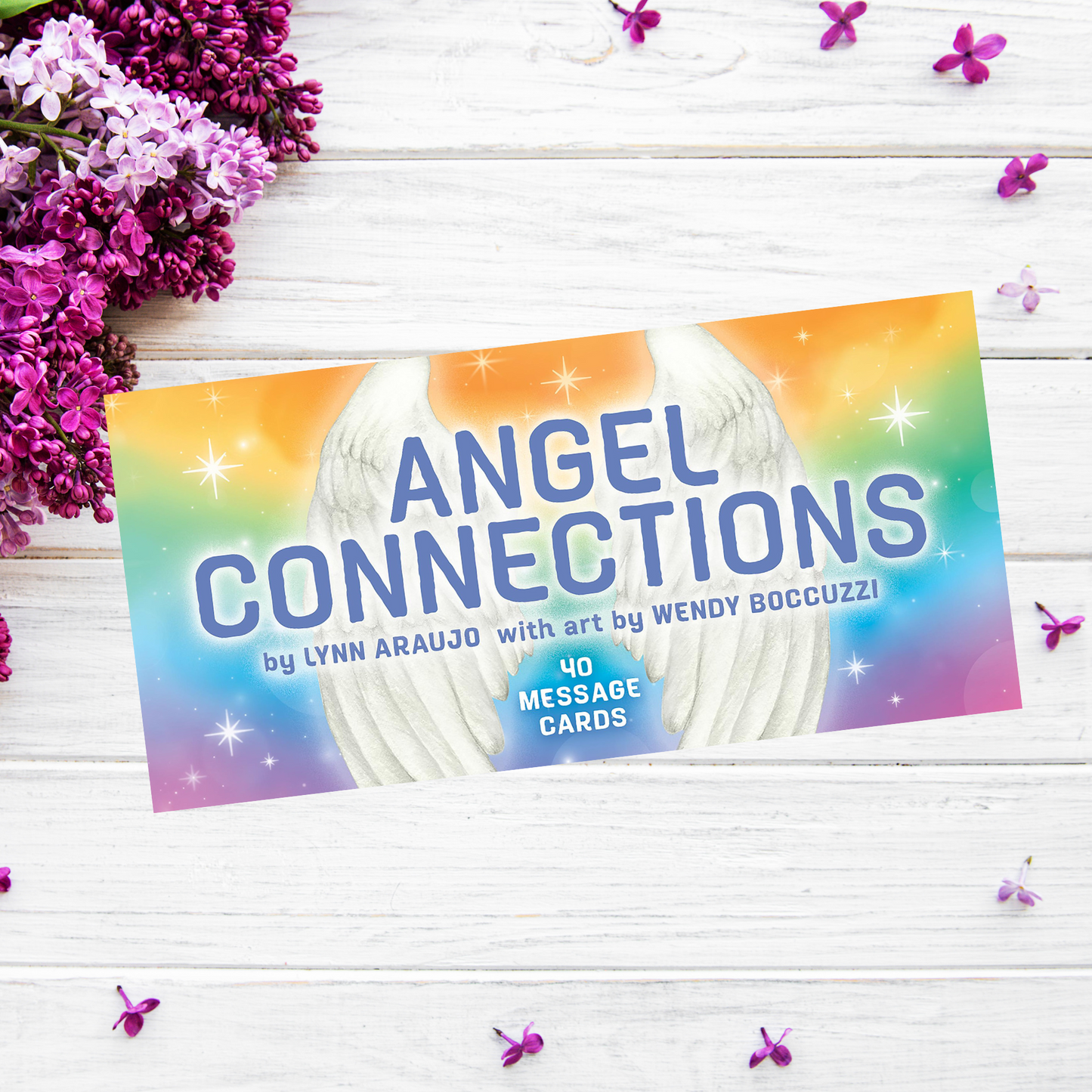 A box labeled "Angel Connections: 40 Message Cards" by Lynn Araujo with art by Wendy Boccuzzi, featuring a bright, colorful background with white angel wings and 40 message cards inside. It rests on a white wooden surface decorated with purple flowers, embodying the essence of angel signs.