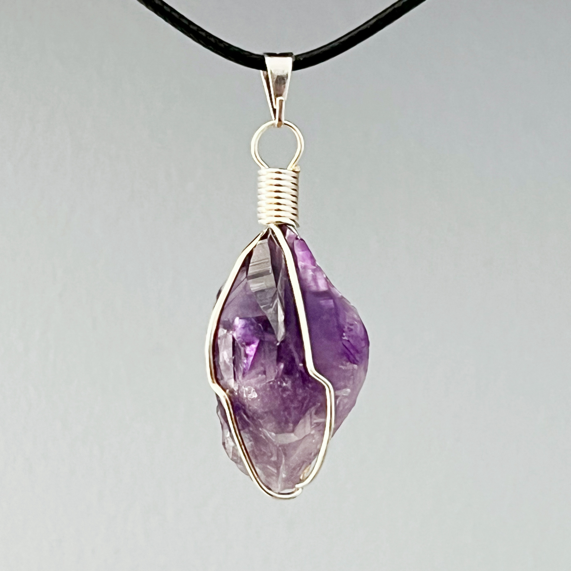 The 1" Wire Wrapped Raw Amethyst Crystal Point Pendant showcases a polished amethyst stone beautifully encased in intricately wrapped silver wire. Suspended on a black cord, the pendant features a purple amethyst with varying shades and visible facets, giving it an authentic, natural appearance. Known for its healing properties, this pendant offers both elegance and charm.