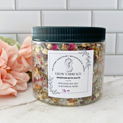 A jar labeled "Eros Embrace Intention Bath Salts" features a luxurious blend of Epsom Salt and Dead Sea Salt, enriched with botanical herbs. A soft pink flower rests beside the jar on the counter, set against white subway tiles. Within the jar is a Rose quartz crystal for added serenity. Net weight: 16 oz.