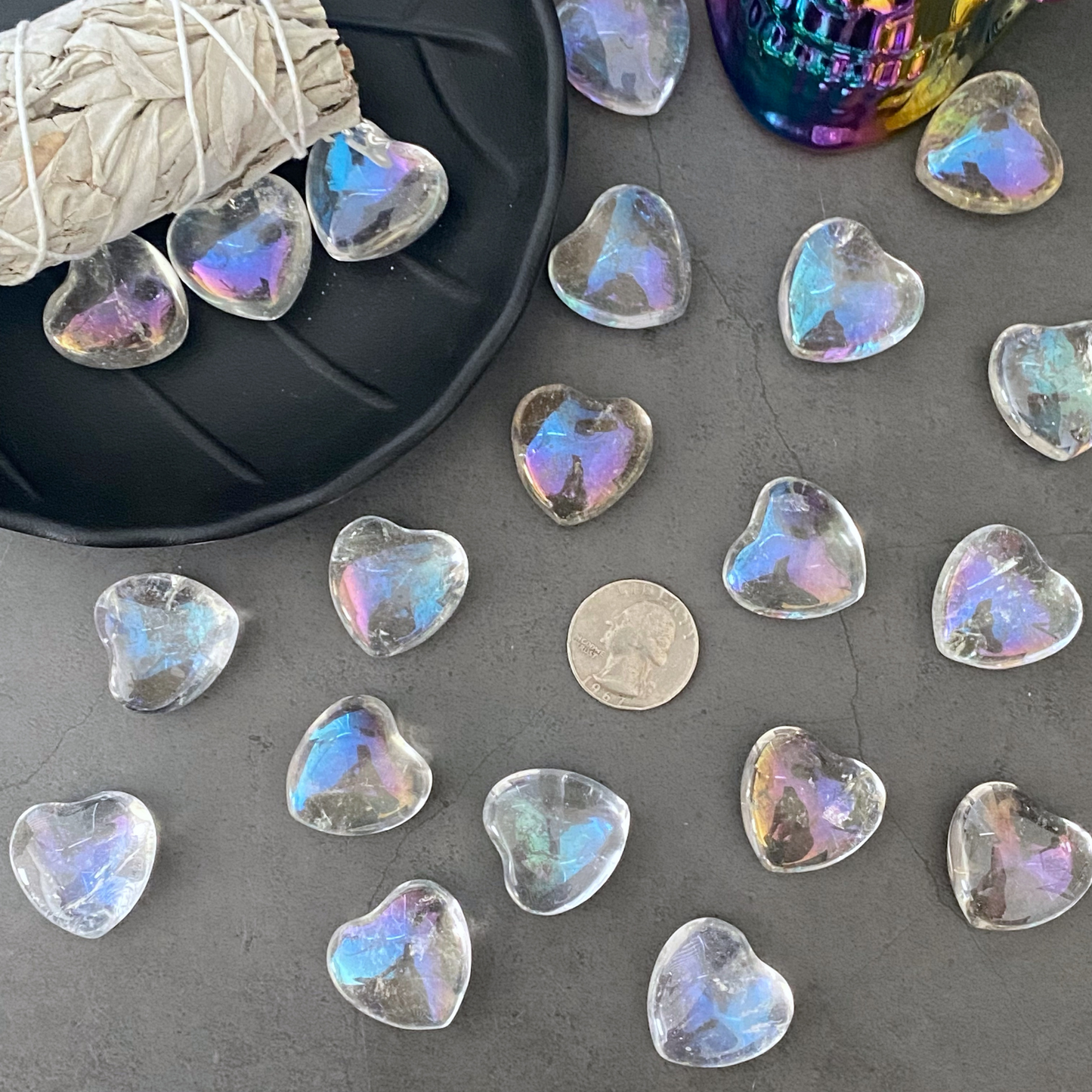 A black dish holds multiple 1" Mini Angel Aura Crystal Quartz Heart stones. The dish is placed on a white marble surface. Nearby, there is a bundle of sage and a colorful, metallic skull.