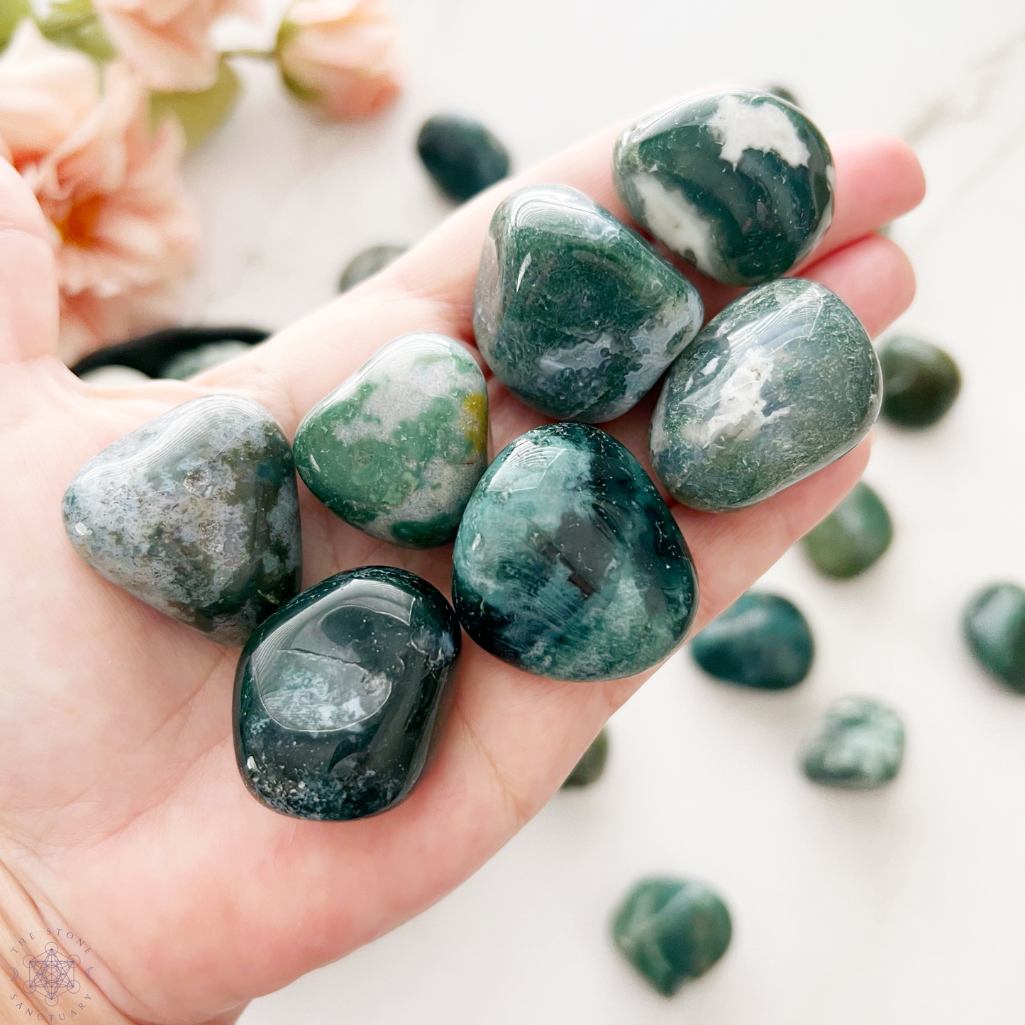 A round wooden bowl filled with polished Green Moss Agate Tumbled Stones of various shades, featuring smooth surfaces and slight color variations. The stones, known for their healing properties and symbolizing new beginnings, are neatly arranged within the bowl, which is set against a plain white background.