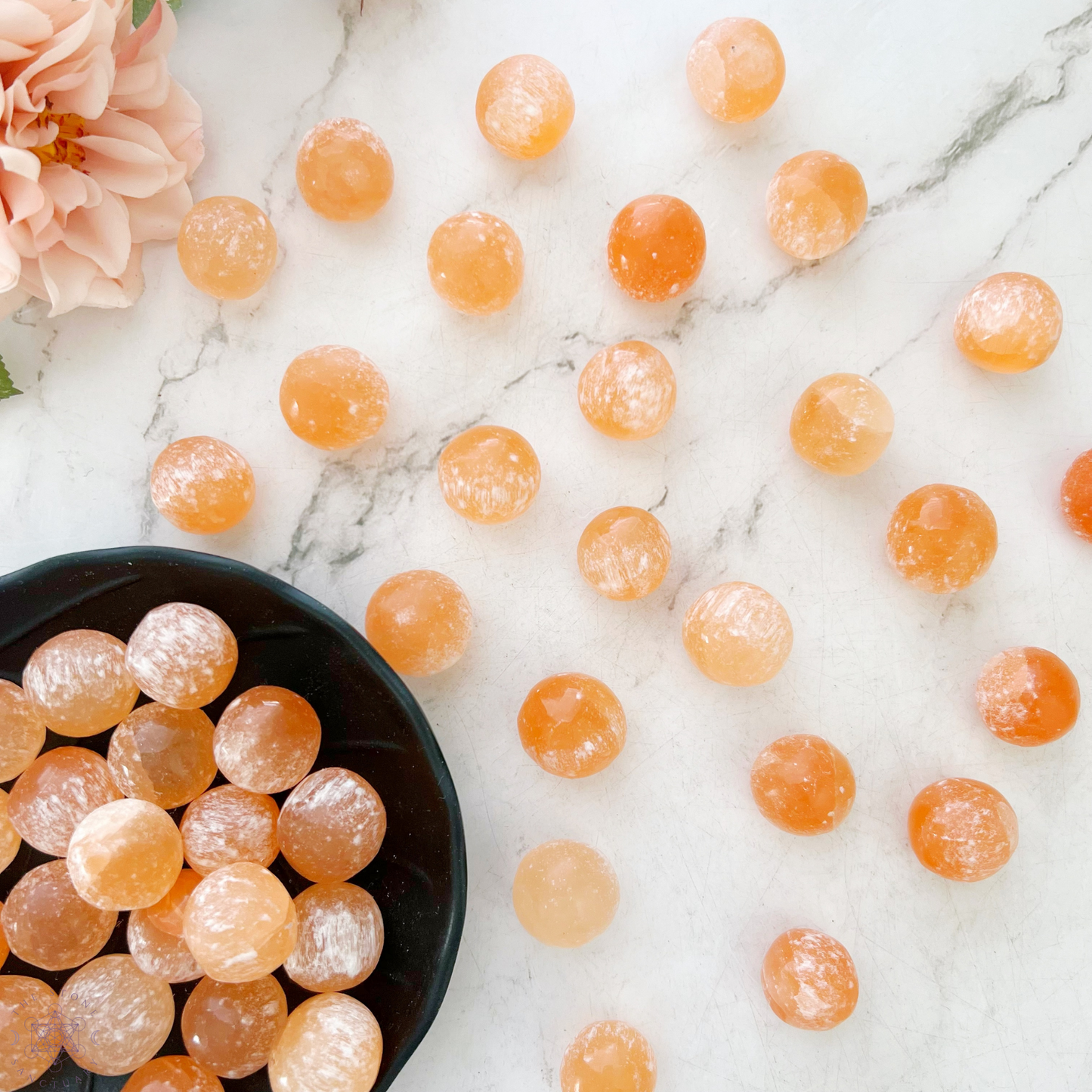 A top view of a wooden bowl filled with numerous round, orange candies resembling Orange Selenite Tumbled Sphere Stones. The candies have a slightly translucent appearance, with a sugary coating visible on their surfaces, invoking thoughts of emotional well-being. The bowl is set against a white background.