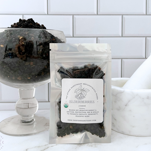 A clear bag labeled "Dried Elderberries" is placed in front of a footed glass bowl filled with dried elderberries. The background features white subway tiles and a white mortar and pestle. The bag has product details, including the botanical name Sambucus nigra, and a website address at the bottom.