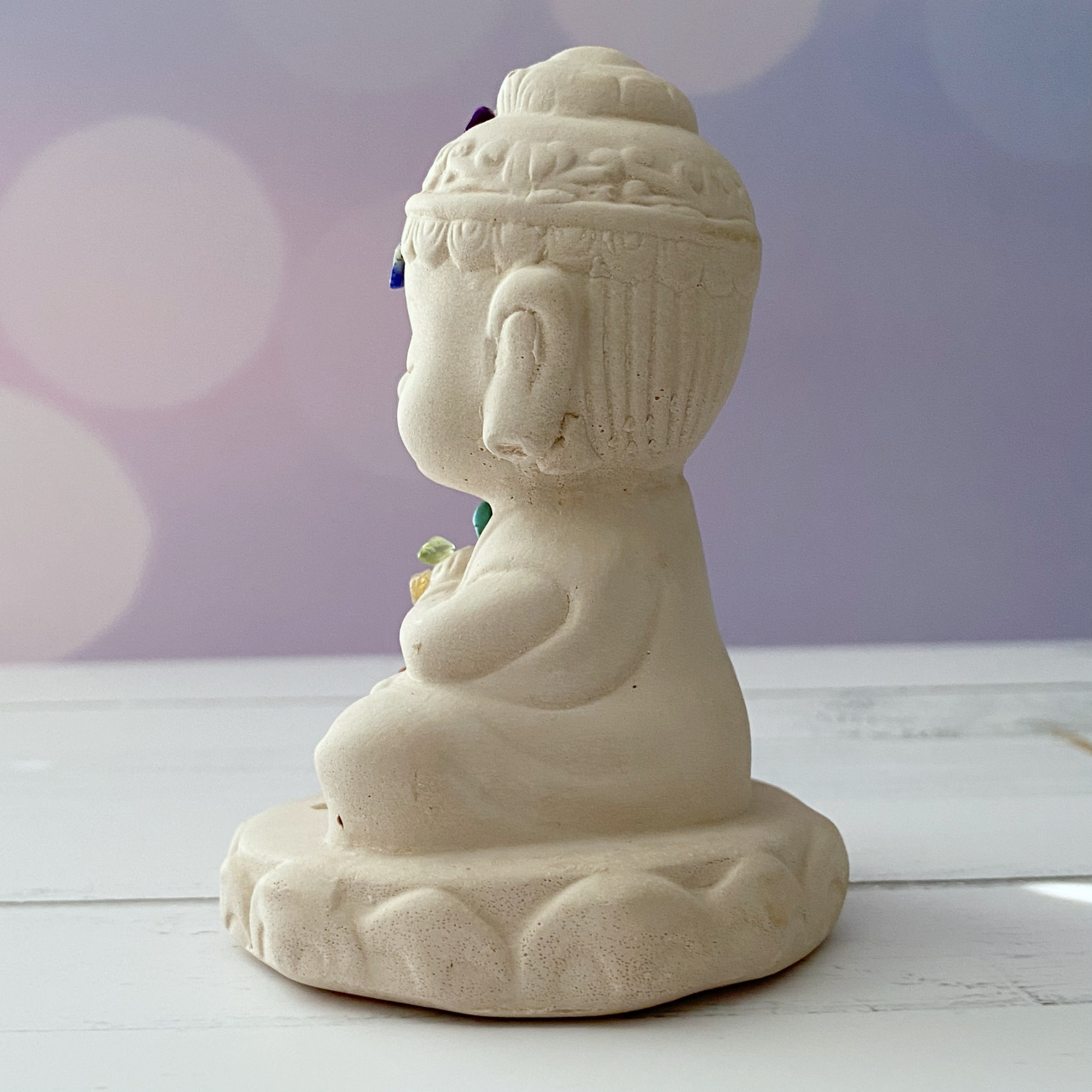 A small ceramic Buddha statue in a seated meditation pose doubles as a Chakra Buddha Incense Holder. The Chakra Buddha Incense Holder features colorful gemstone stone chips on its chakras, representing various energy points. The background is softly blurred with pastel colors and bokeh light effects, set on a white wooden surface.