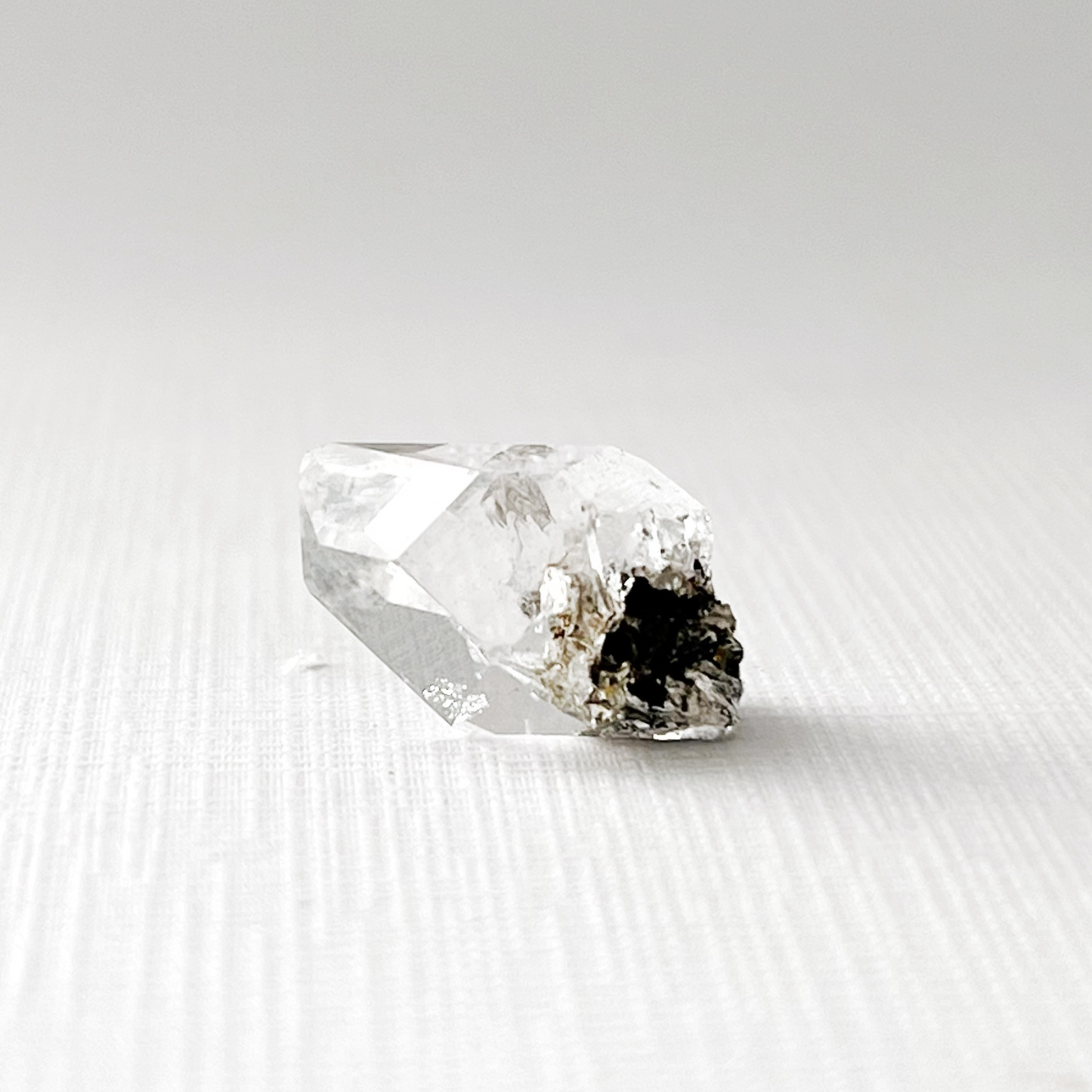 A .63" Raw Herkimer Diamond placed on a reflective black surface creates a mirrored image. The natural quartz crystal catches light, showcasing its translucence and intricate cuts. The background is entirely black, highlighting the brilliance and clarity of these unique formations.