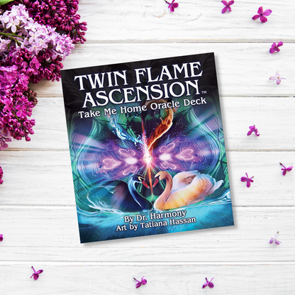 A book titled "Twin Flame Ascension Oracle" is set against a white wooden background with purple flowers scattered around. The cover art by Tatiana Hassan features an ethereal design with two intertwined swans and cosmic elements, embodying the journey of spiritual awakening.