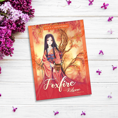 The image shows "Foxfire: The Kitsune Oracle" by Lucy Cavendish and illustrated by Meredith Dillman, lying on a white wooden surface. The cover features an illustrated woman in traditional Japanese attire with a fox by her side, surrounded by decorative purple flowers.