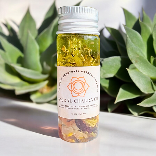 A small glass bottle of Sacral Chakra Oil with yellow liquid and tiny crystals bears a label featuring a geometric symbol and listing benefits like enhanced creativity, emotional balance, and improved relationships. The bottle is nestled amid green succulent plants.