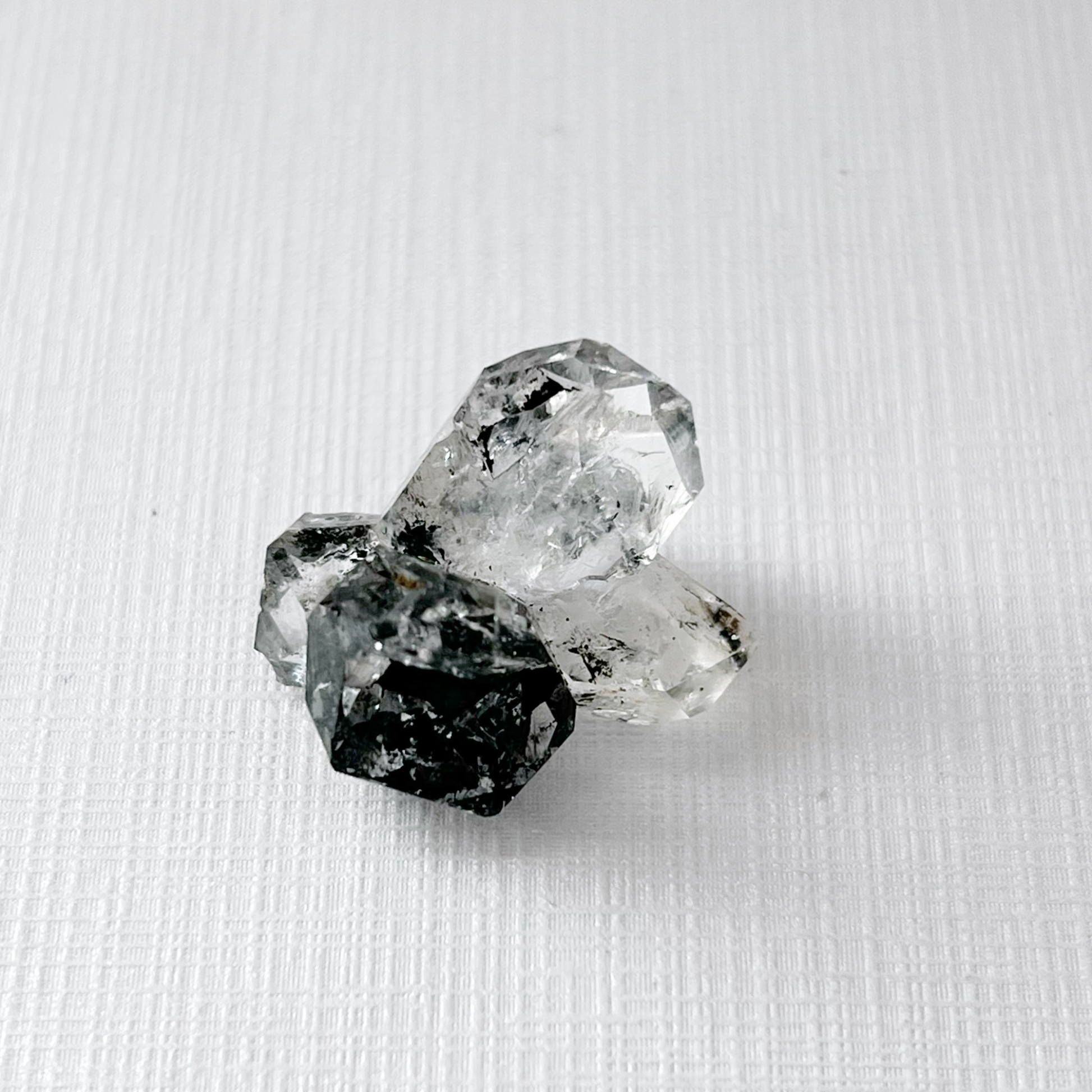 A close-up image of three clear, reflective, irregularly-shaped crystals placed on a glossy black surface, creating a striking reflection below them. The dark background enhances the brilliance and clarity of this .72" Raw Herkimer Diamond Cluster with Hydrocarbon Anthraxolite, revealing its metaphysical properties.