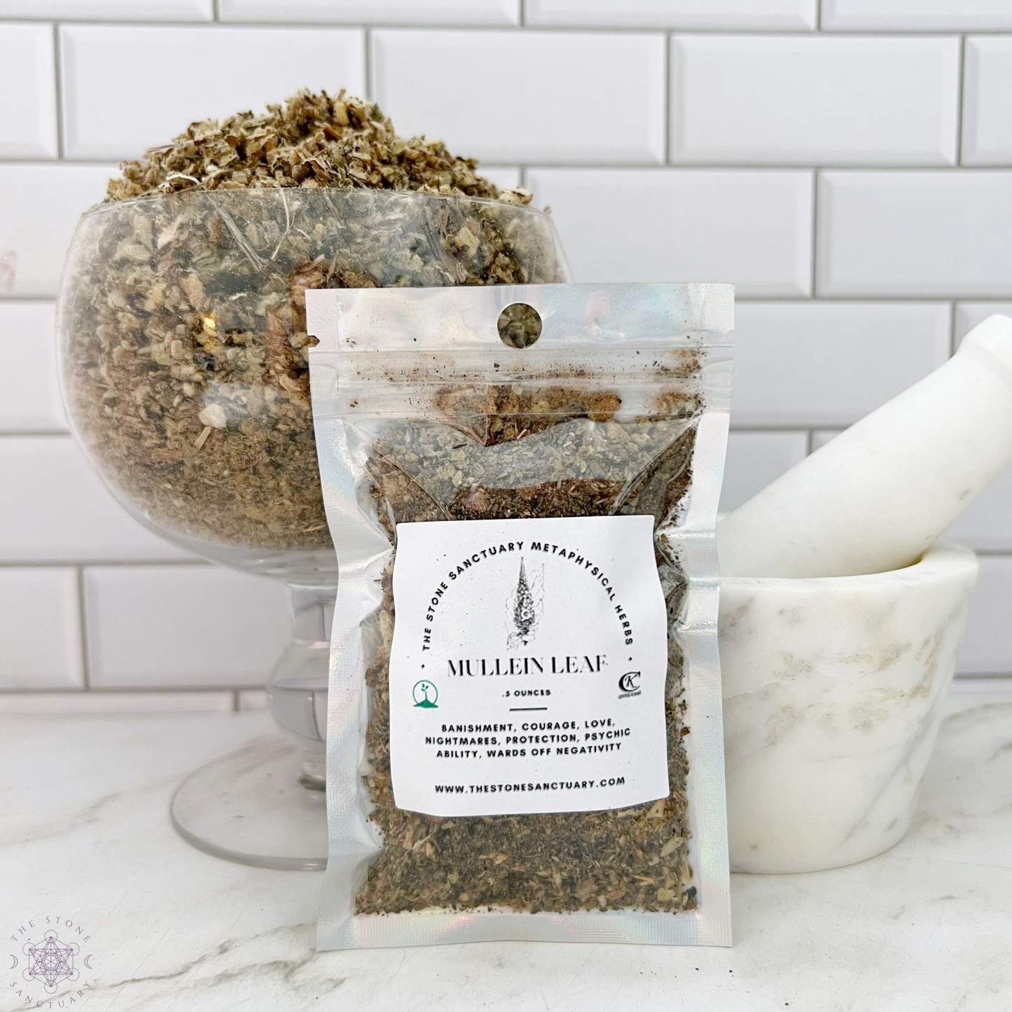 A glass container filled with wildcrafted USA dried Mullein Leaf sits on a counter next to a white mortar and pestle. In front of the container is a small, sealed packet of Mullein Leaf with a label featuring product information against a white tile background.