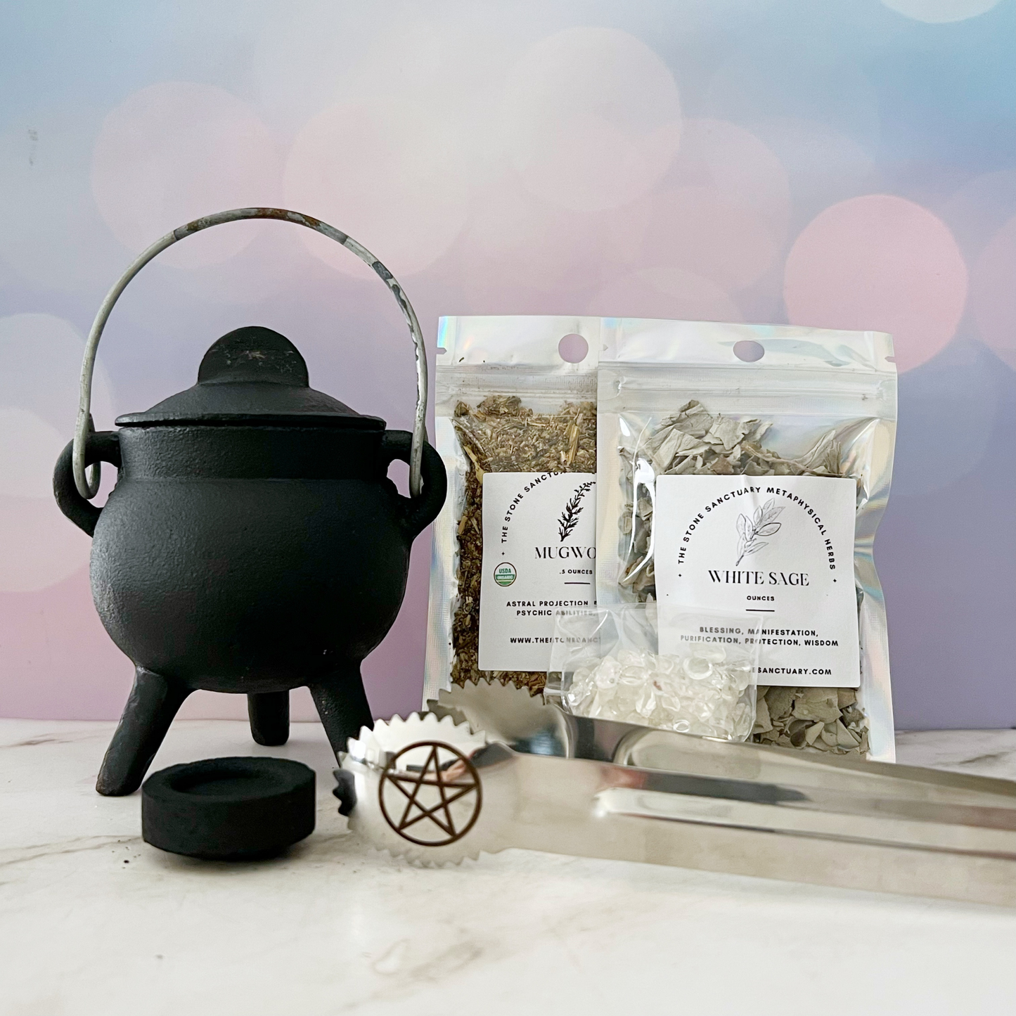 A compact Mini Cast Iron Cauldron sits on a table alongside two bags of herbs labeled "Mugwort" and "White Sage." In front, there are a pair of metal tongs adorned with a pentagram symbol, crystal fragments, and a round charcoal disc. This Mini Cast Iron Cauldron Kit creates an inviting atmosphere for your spiritual practice.