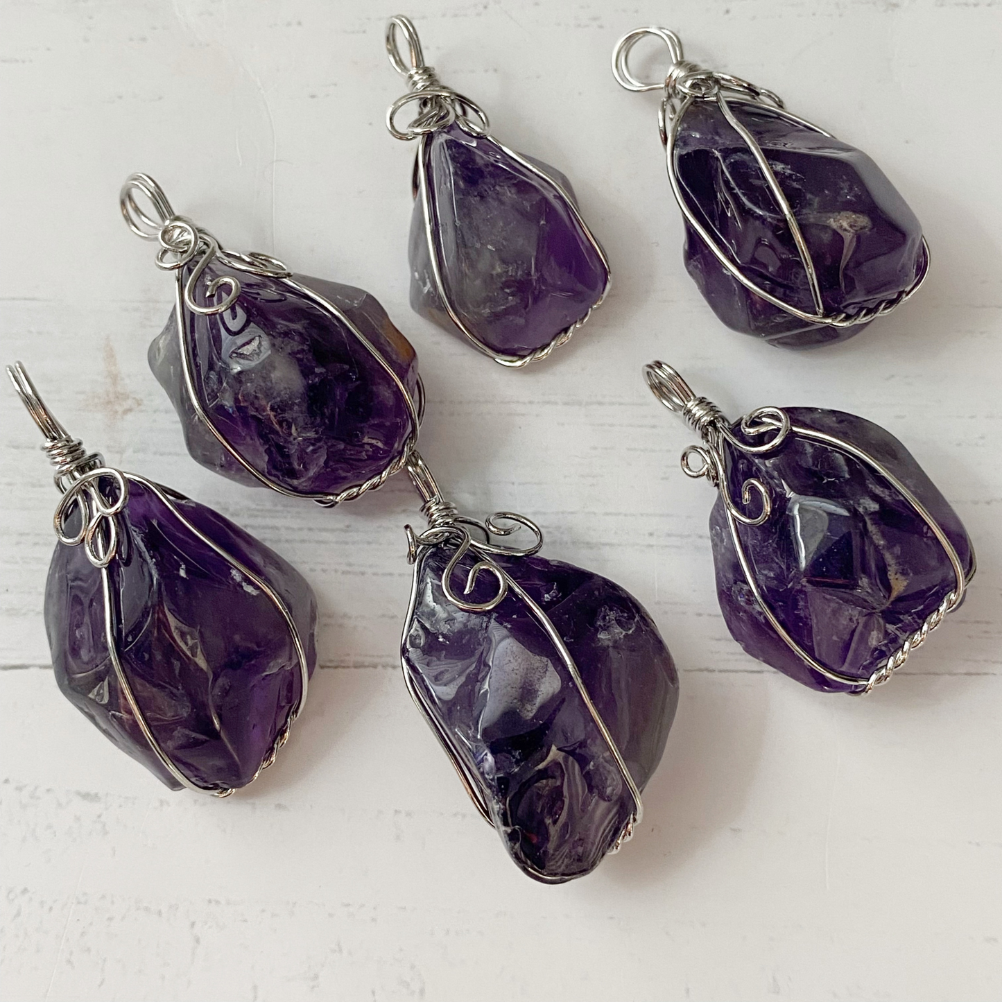 A close-up of a 1.25" Wire Wrapped Rough Amethyst Crystal Pendant wrapped in intricate silver wirework, hanging on a black cord. The crystal displays varying shades of purple. The blurred, neutral-toned background highlights the pendant's details. This piece not only enhances spiritual awareness but also wards off negative energies.